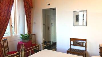 Habitación de hotel con una cama, una silla de madera, una mesita con una planta, una silla roja, una ventana con cortinas y una puerta que da a otra habitación.