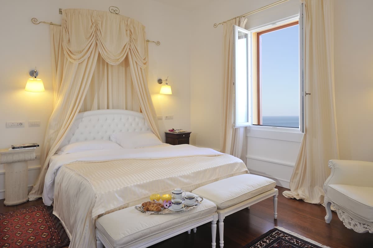 Un dormitorio con una cama grande, una mesita de noche, un banco y una ventana con cortinas.