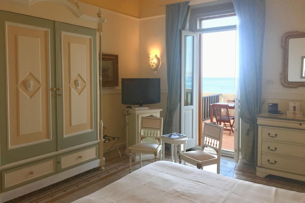 Habitación de hotel con una cama grande, televisión, escritorio con silla y vista al mar a través de una puerta de balcón.
