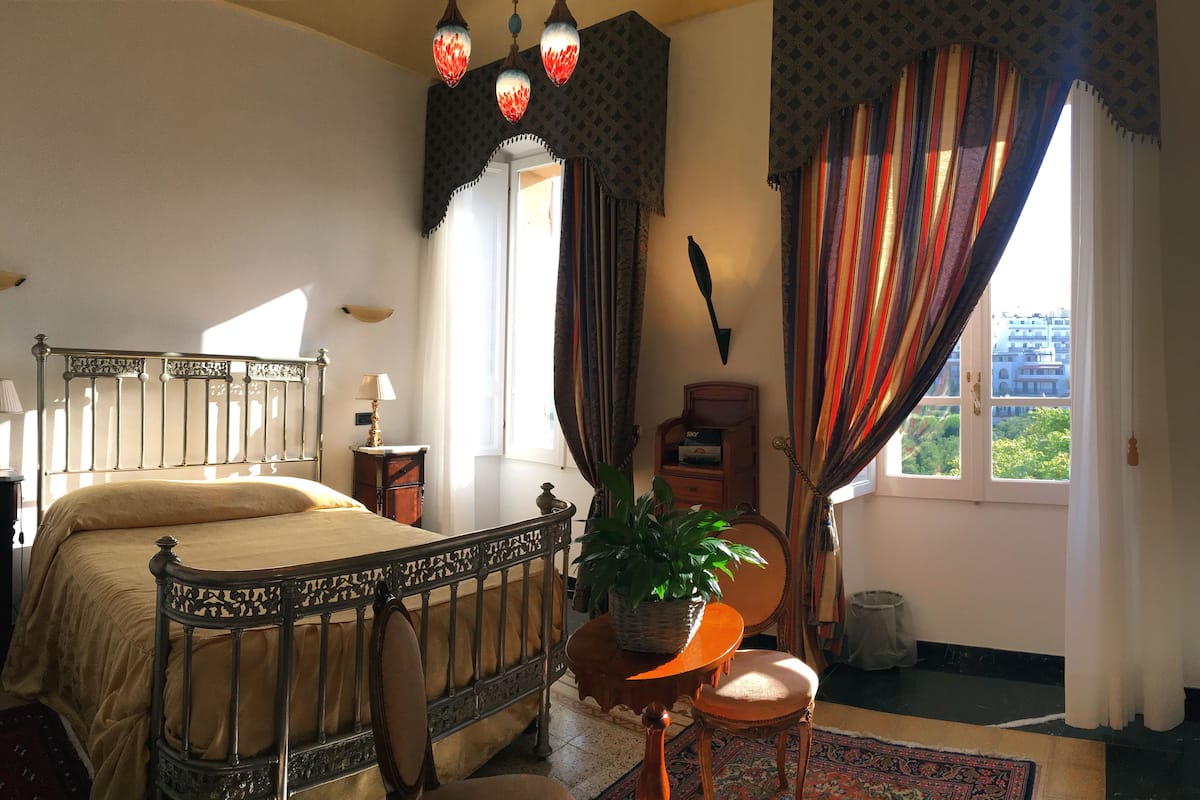 Un dormitorio con cama, mesita de noche, lámpara, planta en maceta, silla, ventana con cortinas y vista a follaje exterior.