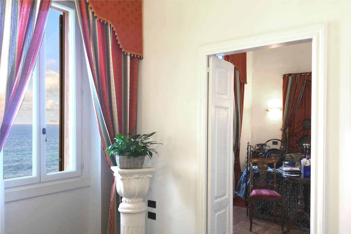 Una habitación con vista al mar a través de una ventana, una planta en maceta sobre un pedestal, y un comedor con una silla y una mesa.