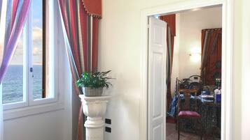 Une pièce avec vue sur la mer, visible par une fenêtre, un pot de fleurs posé sur un piédestal, et un coin repas avec une chaise et une table.