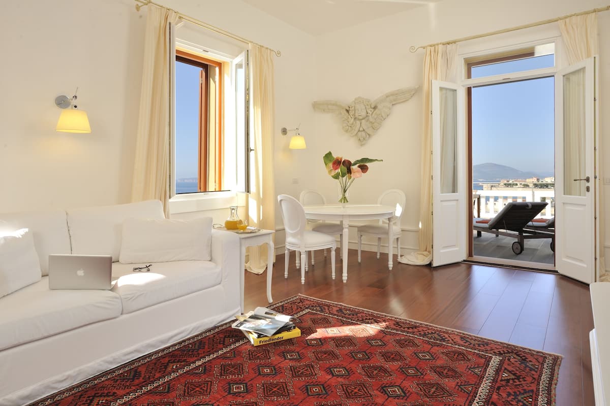 Una sala moderna con un sofá blanco, una mesa de comedor con sillas, un tapete rojo estampado y vista al mar a través de una puerta abierta.