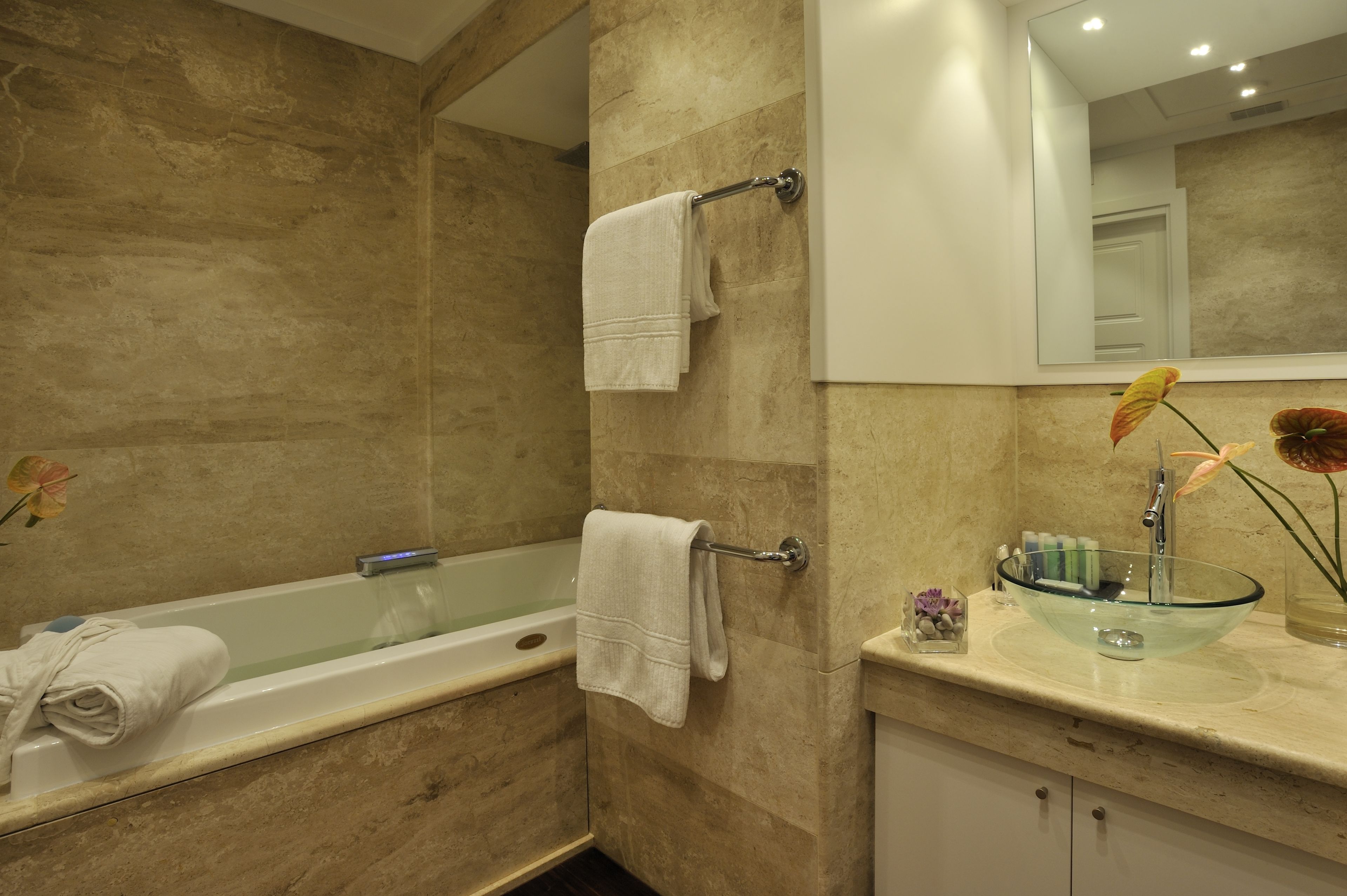 Un bagno con una vasca, due asciugamani su porta-asciugamani, un lavandino in vetro con un vaso di fiori e uno specchio sopra il lavandino.