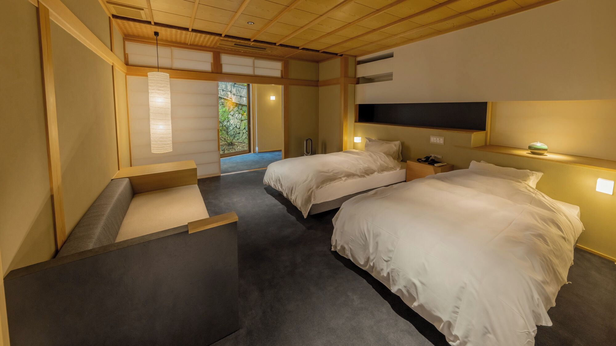 A hotel room with two beds, a wooden ceiling, a view of greenery, and a small table with a lamp.