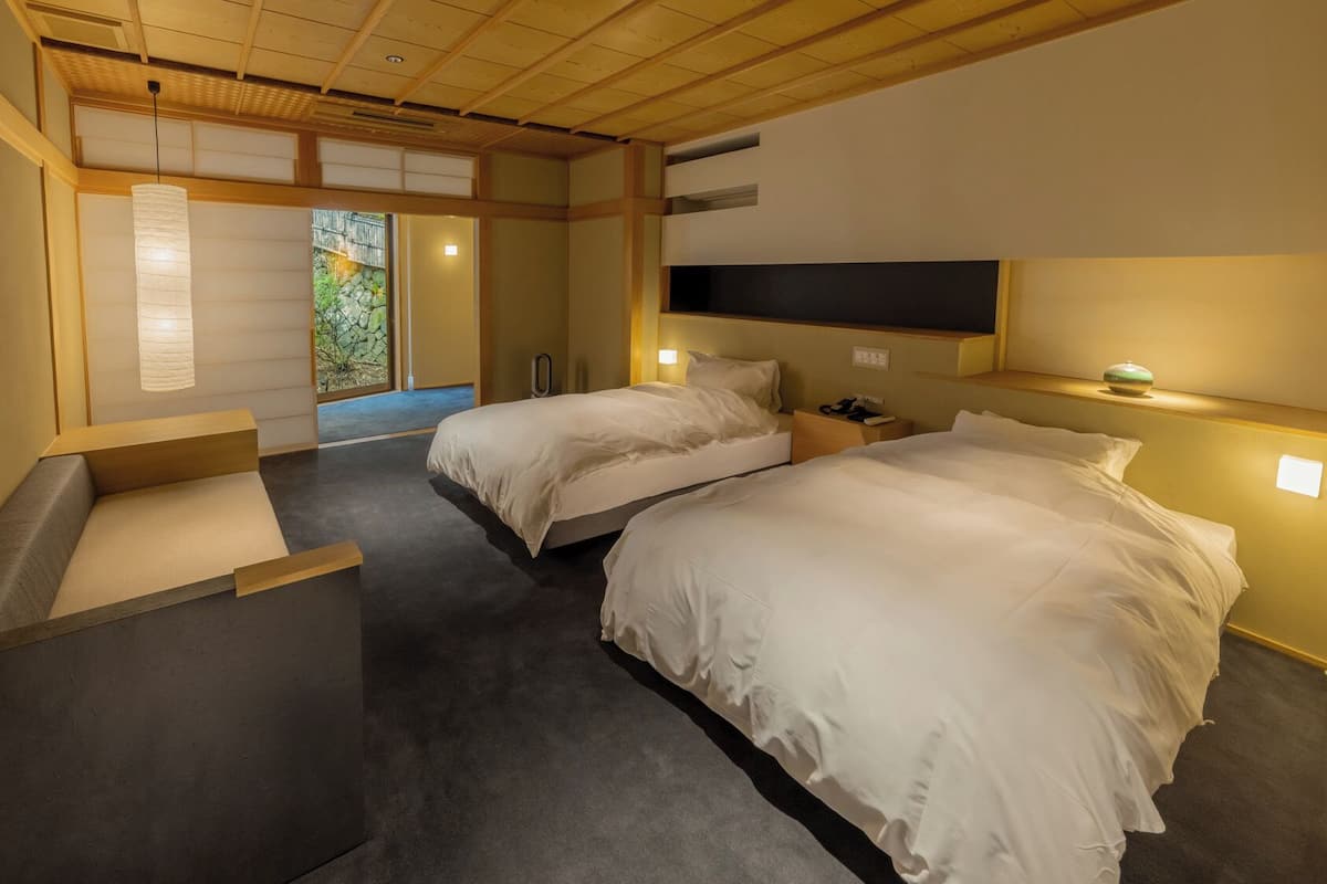 A hotel room with two beds, a wooden ceiling, a view of greenery, and a small table with a lamp.