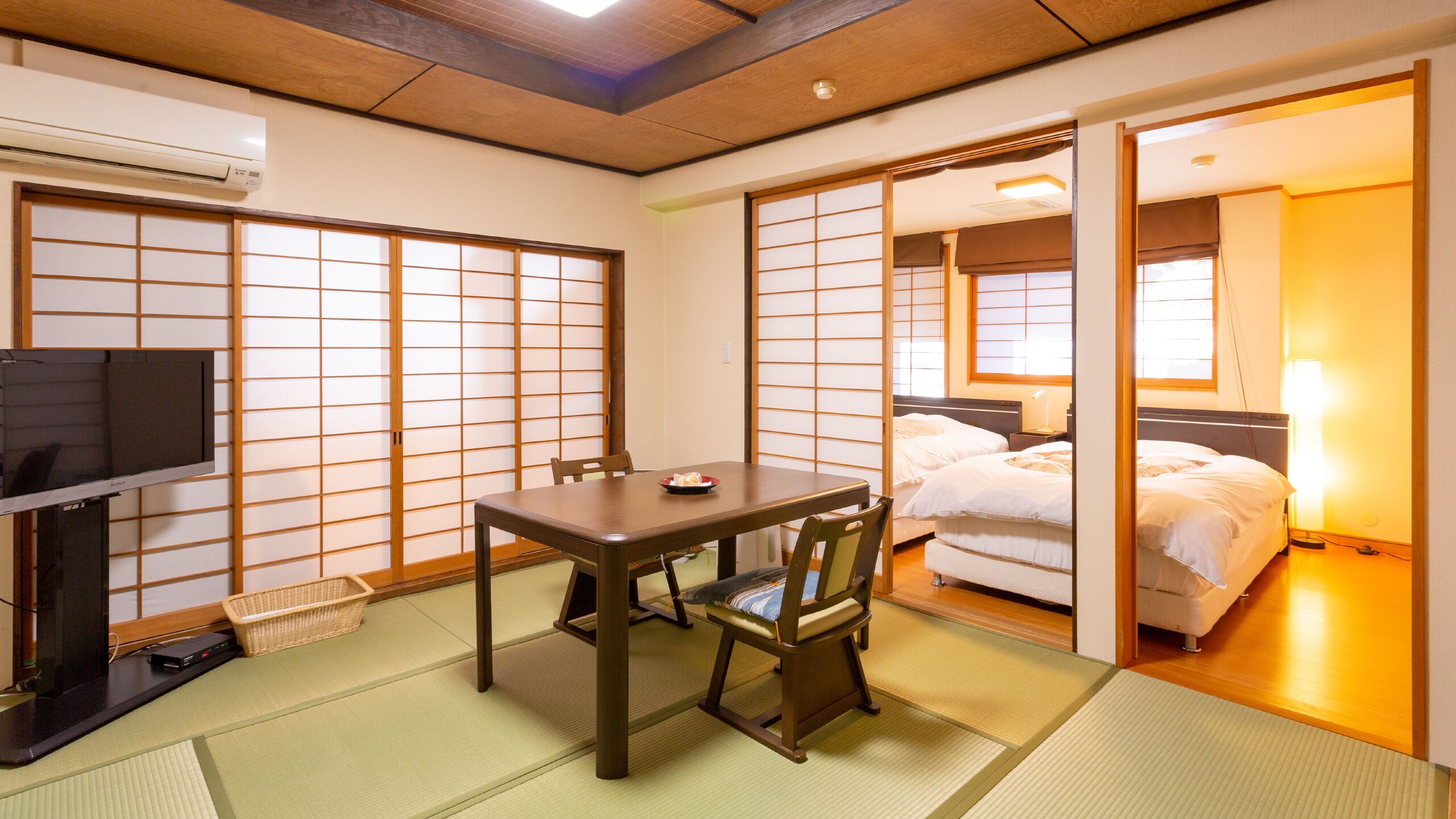 A traditional Japanese room with tatami flooring, a low table, a TV, and a bed with a nightstand.