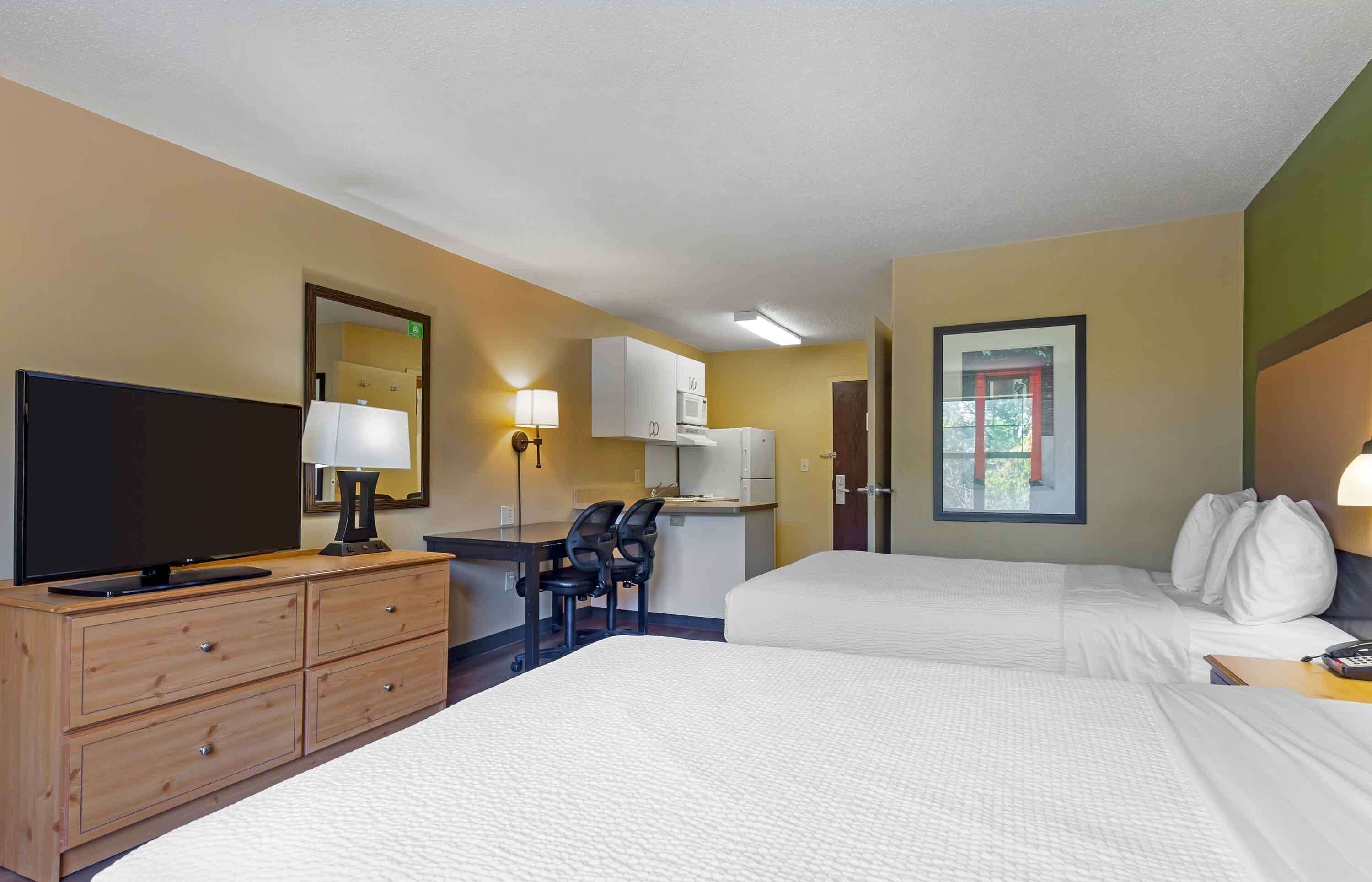 A hotel room with two beds, a desk with a TV, a chair, a desk lamp, a microwave, and a window with a view of trees.