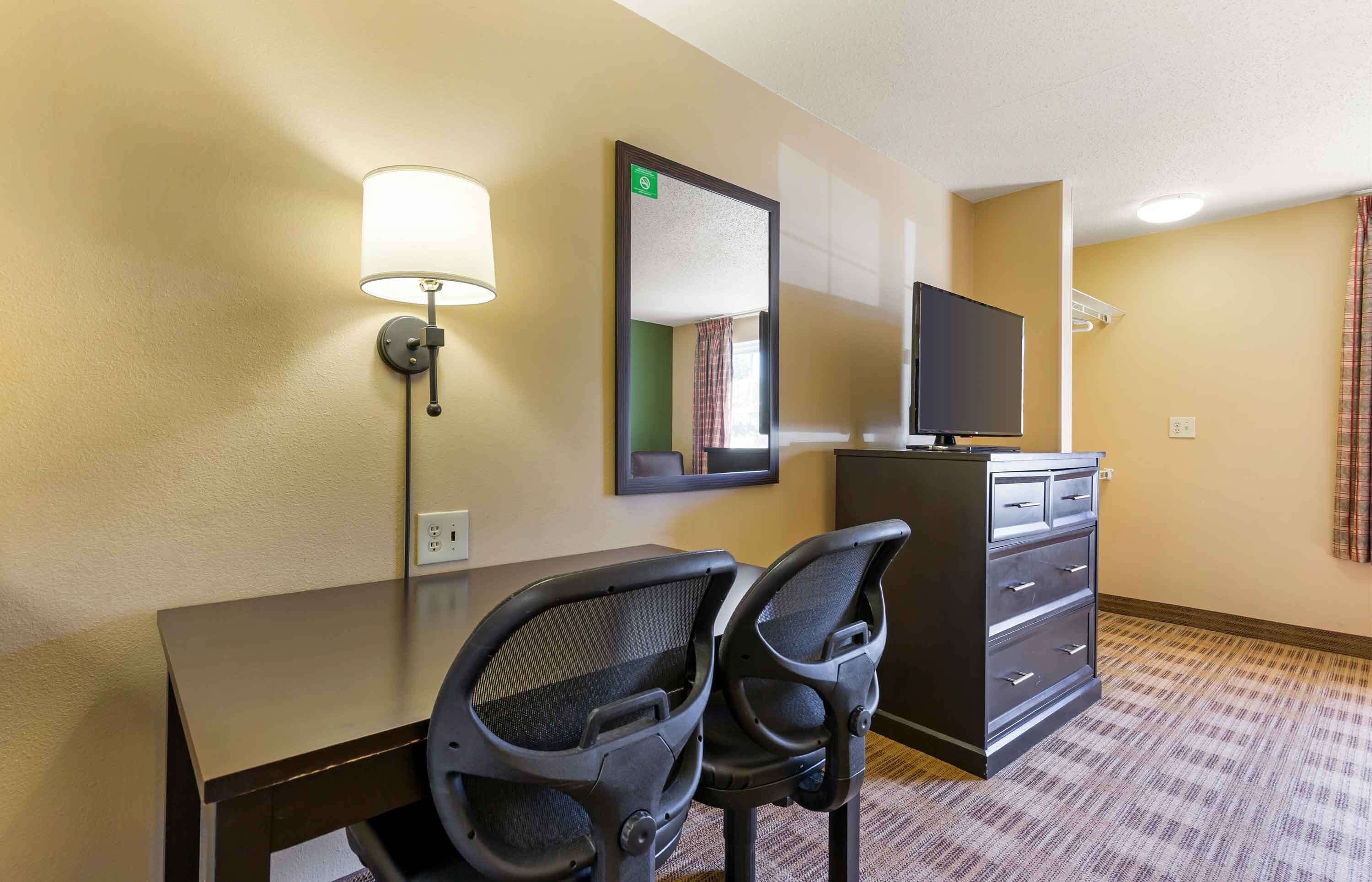 Habitación de hotel con escritorio, dos sillas de oficina, televisor, espejo y cómoda.