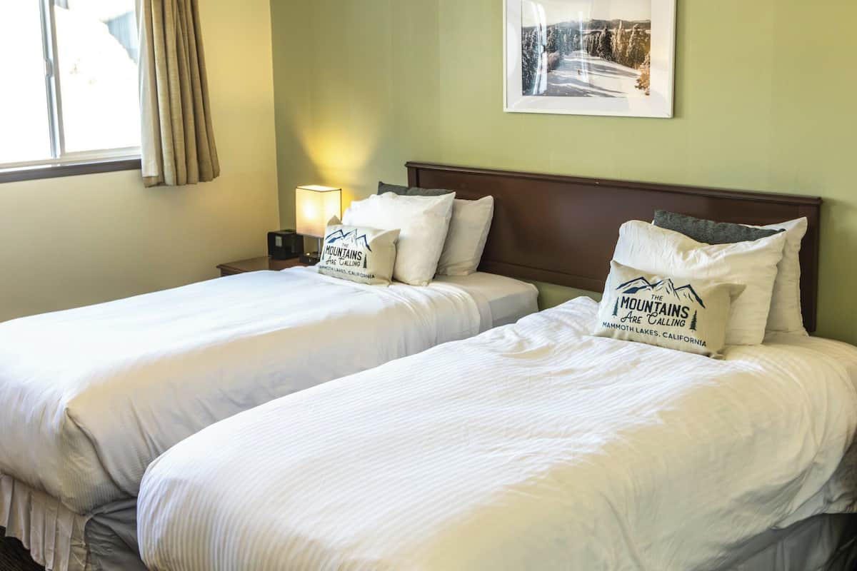 A hotel room with two beds, a nightstand with a lamp, a framed picture on the wall, and a window with curtains.