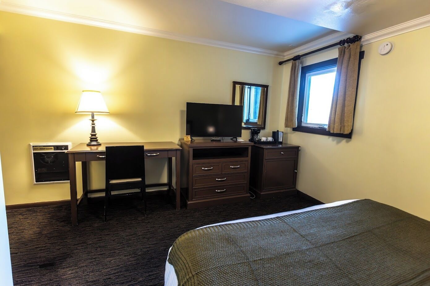 Habitación de hotel con cama, escritorio con lámpara, televisión, cómoda y ventana con cortinas.
