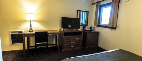 Habitación de hotel con cama, escritorio con lámpara, televisión, cómoda y ventana con cortinas.