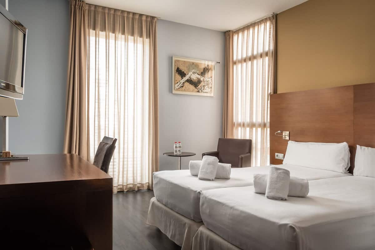 A hotel room with two beds, a desk, a chair, a small table with water bottles, and a painting on the wall.