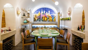 A dining area with a round glass table set for six, surrounded by wooden chairs. A wine rack is visible to the left, and a colorful mosaic wall is behind the table. - Hotel Meninas (Madrid)
