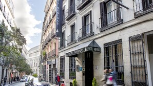 A street view of Hotel Meninas with a cobblestone road, parked cars, and pedestrians. - Hotel Meninas (Madrid)