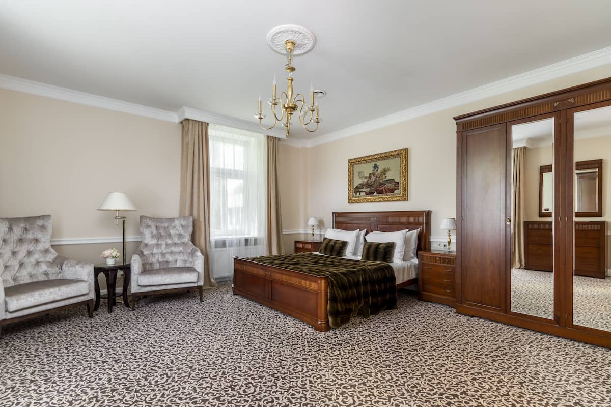 A hotel room with a large bed, two armchairs, a nightstand, a lamp, a chandelier, a mirror, and a patterned carpet.