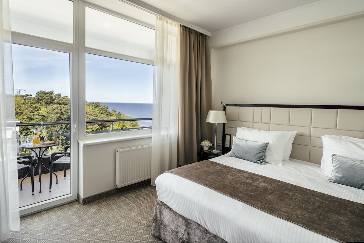 A hotel room with a large window, a bed with white linens, a brown throw, and a nightstand with a lamp.