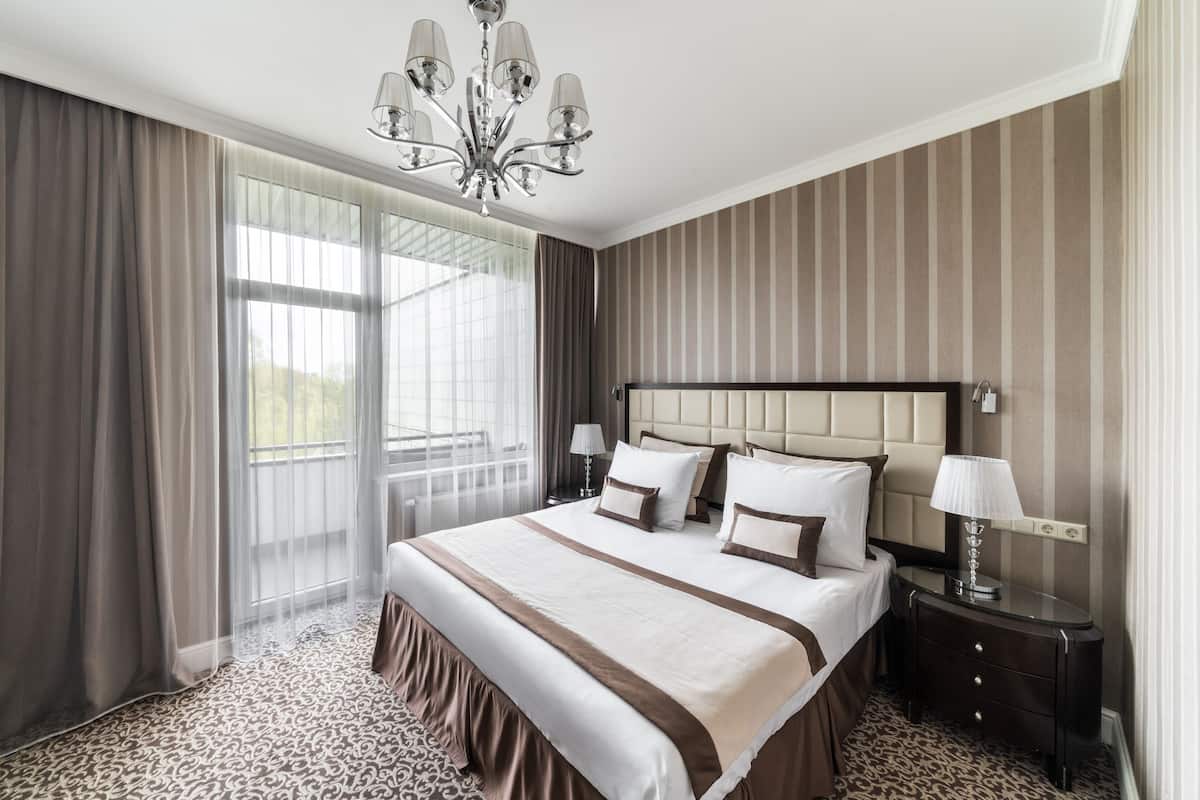 A hotel room with a large bed, a chandelier, a nightstand, and a window with curtains.