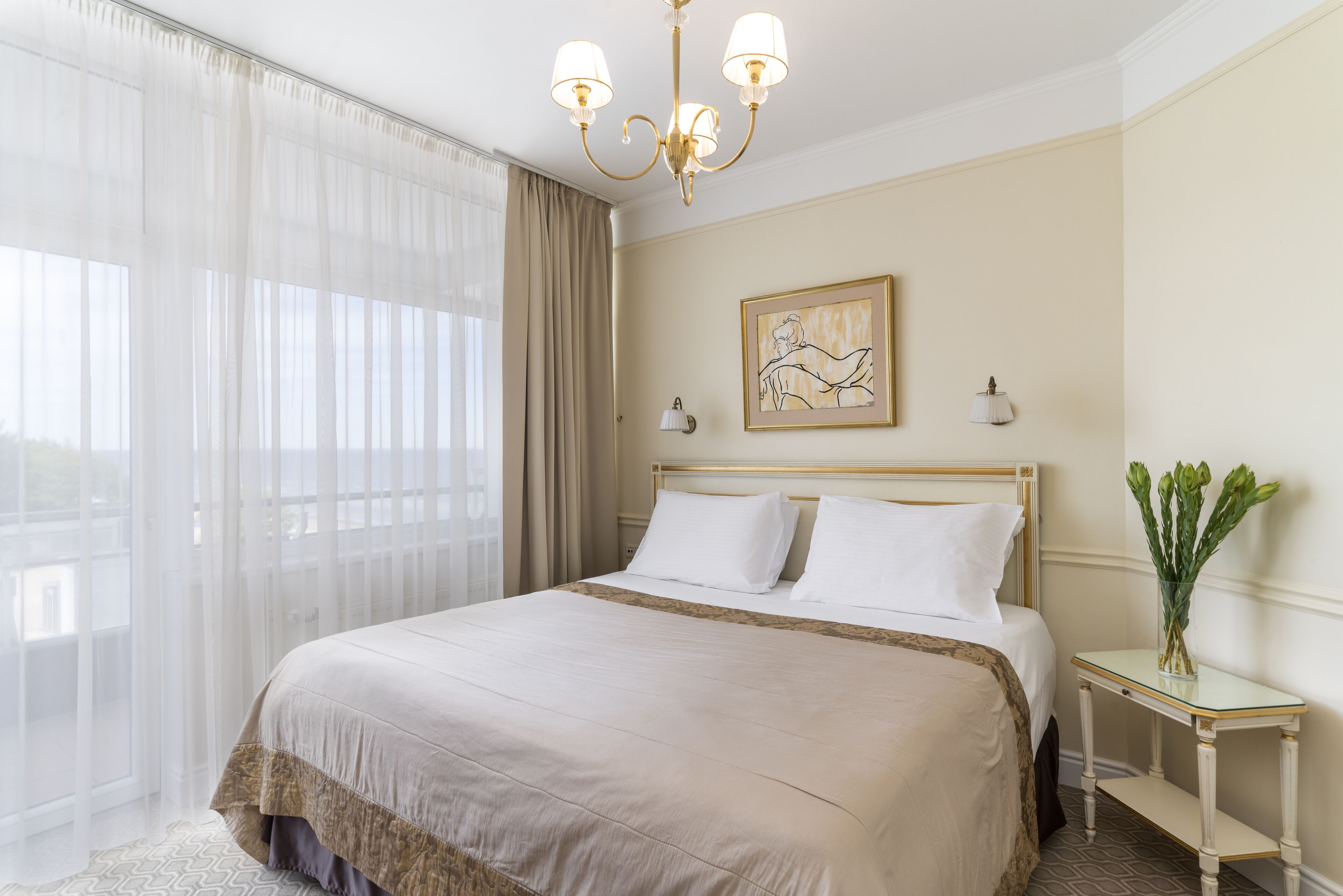 Habitación de hotel con una cama grande, un candelabro, un cuadro en la pared y un florero con flores sobre una mesita.