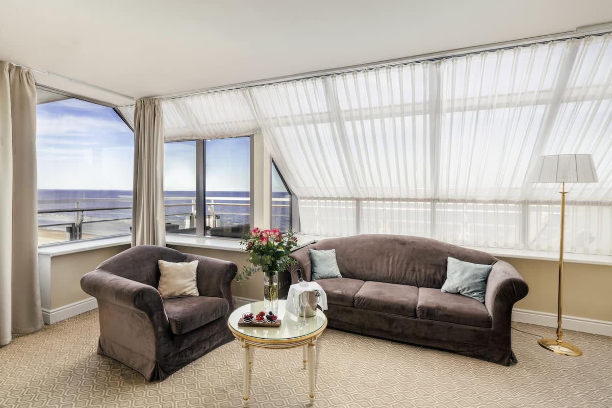A room with a brown sofa, a small round table with a vase of flowers, and a view of the sea through large windows.