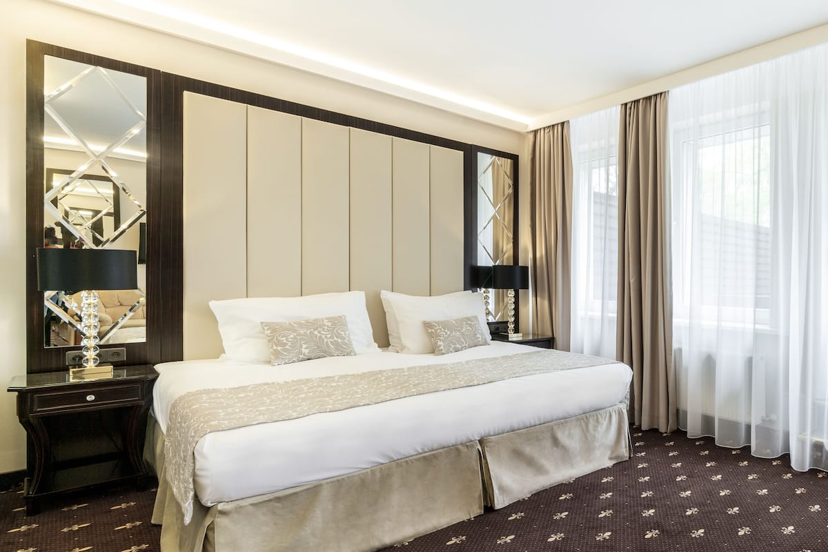 A hotel room with a large bed, a nightstand with a lamp, a mirror, and a window with curtains.