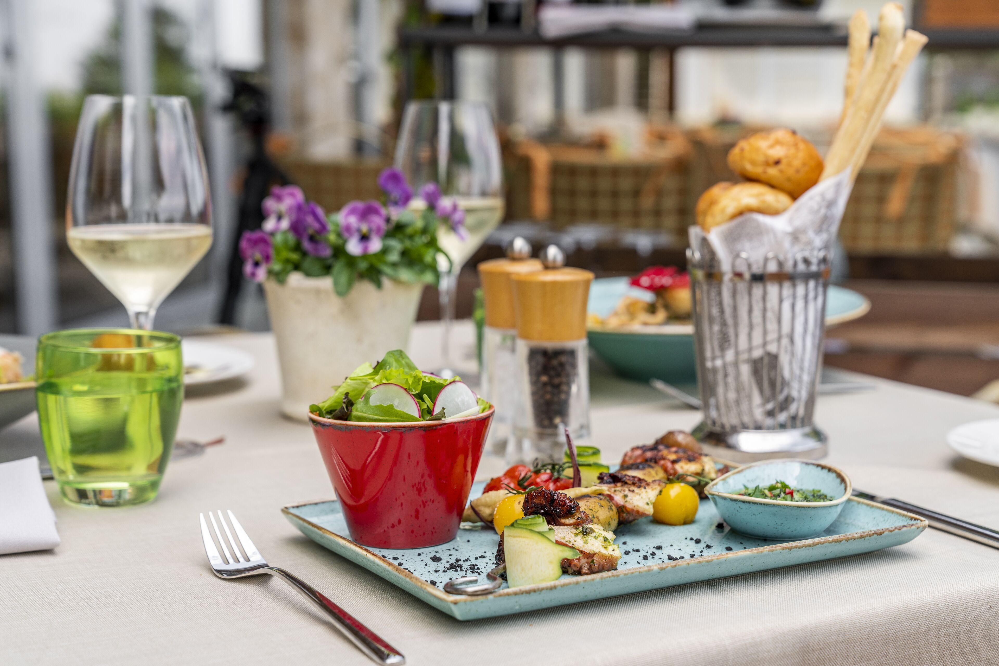 A dining table with a variety of dishes, including a salad, grilled meat, and bread. There are also glasses of wine and a vase with flowers.
