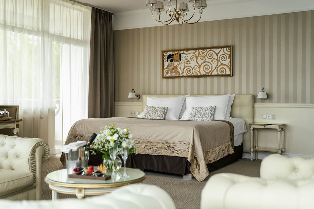 A hotel room with a large bed, a bedside table with a lamp, a decorative wall piece, and a view of the outdoors through sheer curtains.