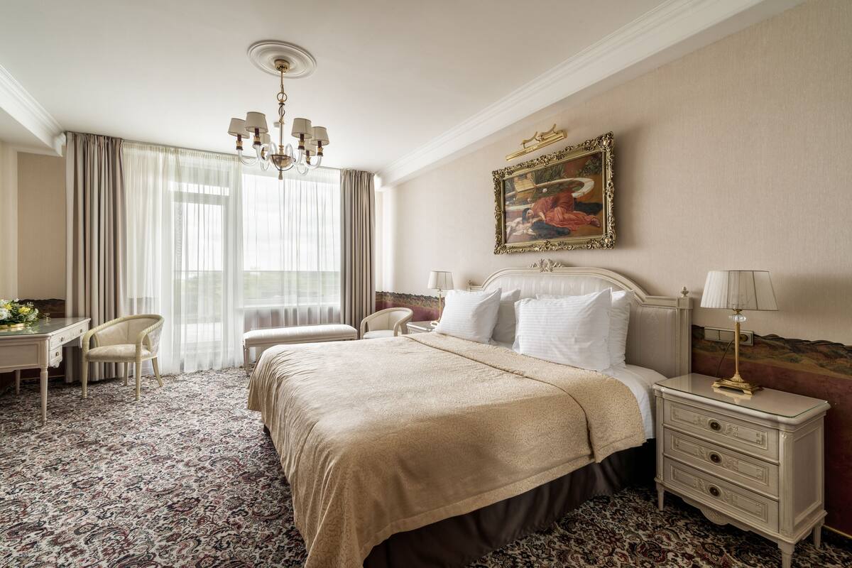 A hotel room with a large bed, a desk, a chair, and a nightstand with a lamp.