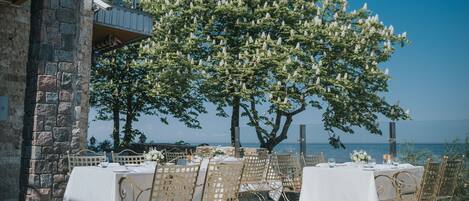Un espacio de comedor al aire libre con mesas y sillas, un muro de piedra y un árbol con flores blancas.