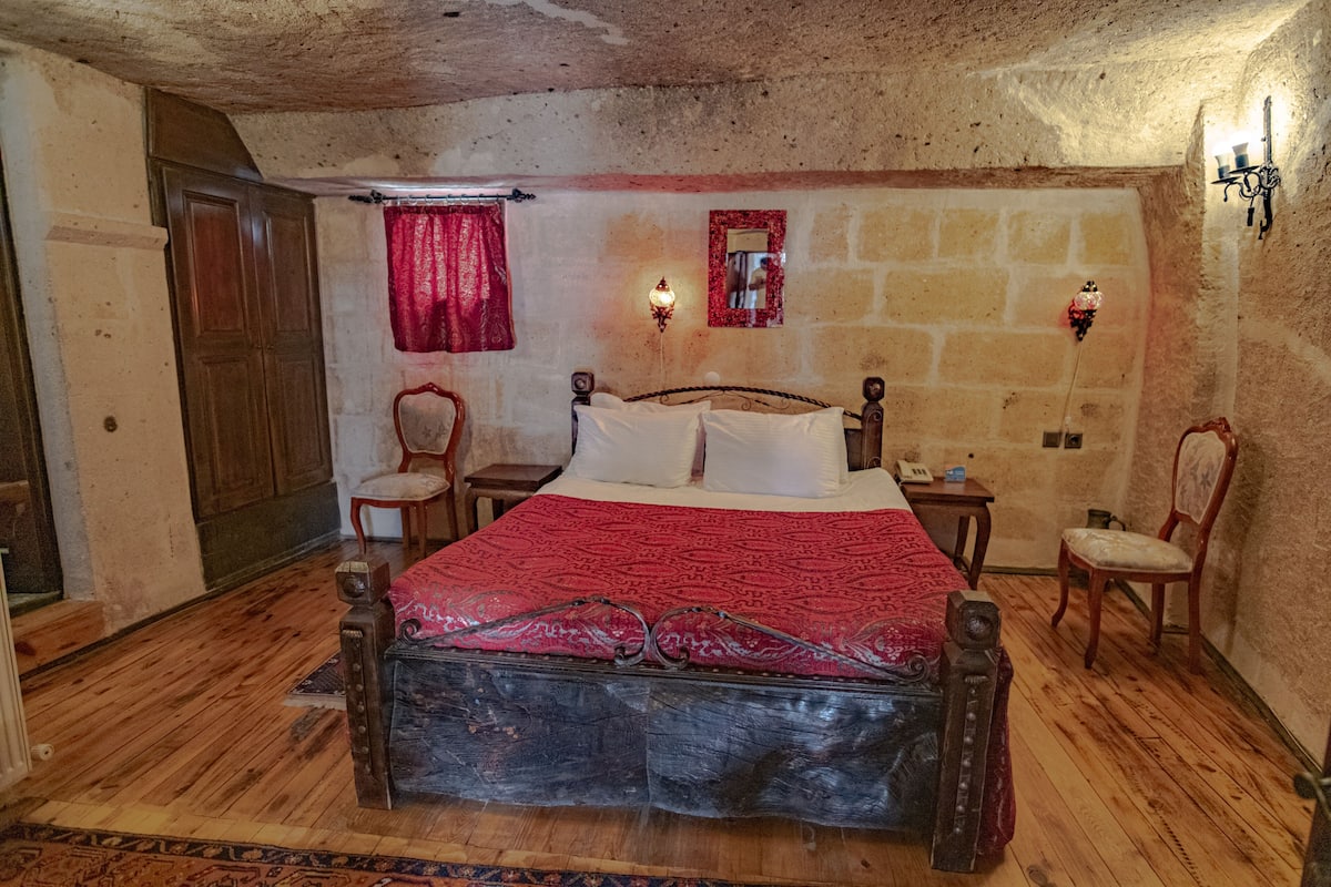 Quarto rústico com cama de madeira, duas cadeiras, mesa de cabeceira e paredes de pedra.