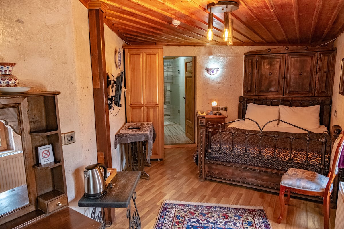 Um quarto aconchegante com teto de madeira, uma cama com colcha estampada, uma cadeira, uma mesa pequena com chaleira e um tapete no chão.