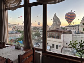 Vista desde una ventana sobre un paisaje urbano con globos aerostáticos, una destacada formación rocosa y edificios con antenas parabólicas.