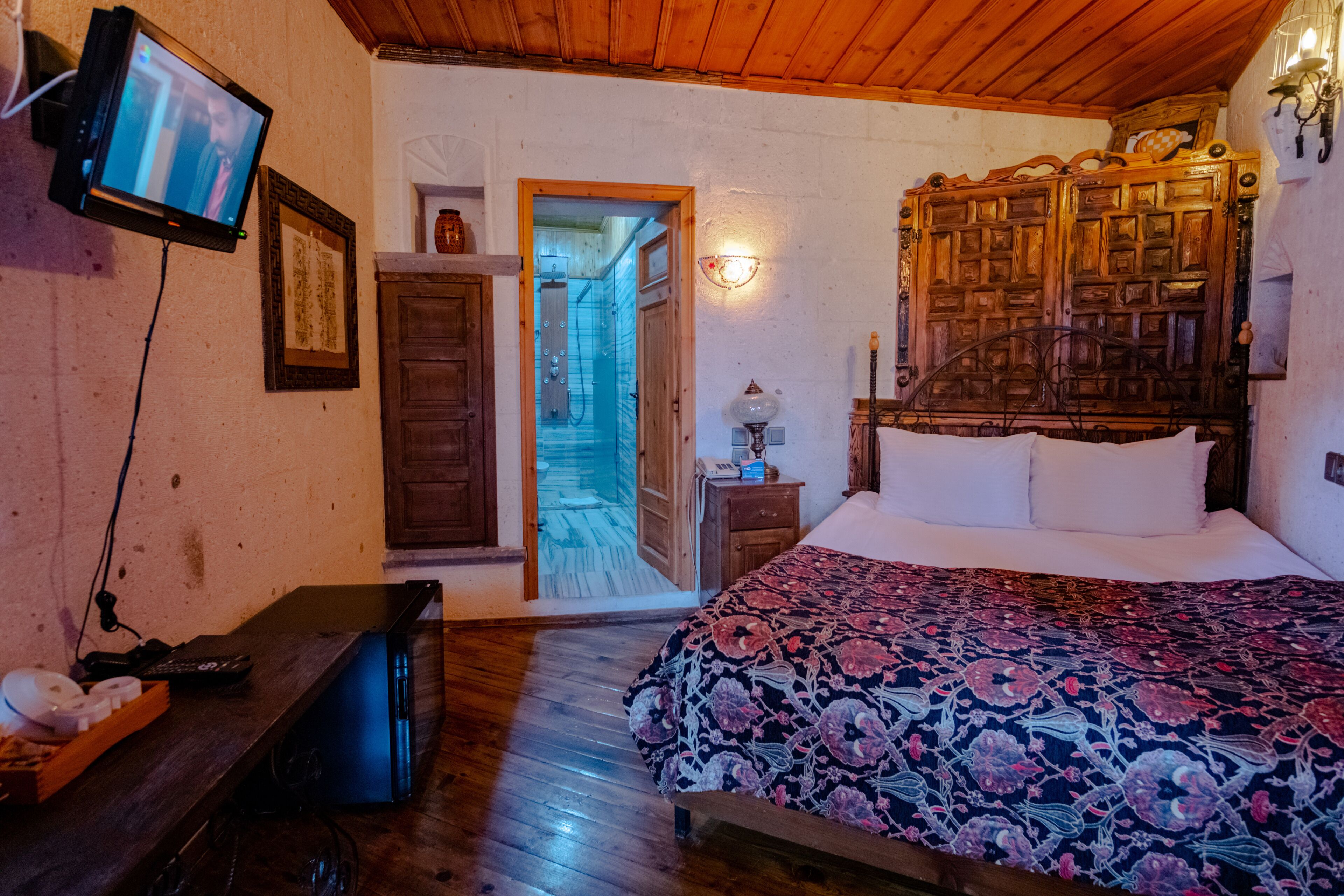 Un dormitorio con cabecera de madera, una cama con colcha floral, un televisor montado en la pared y un aparador de madera.