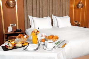 Una cama bien hecha con sábanas blancas, una bandeja de desayuno con pasteles, frutas y una taza de café.