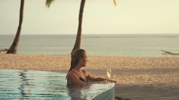 Una persona relajándose en una piscina, con una bebida, con vistas a una playa y palmeras.