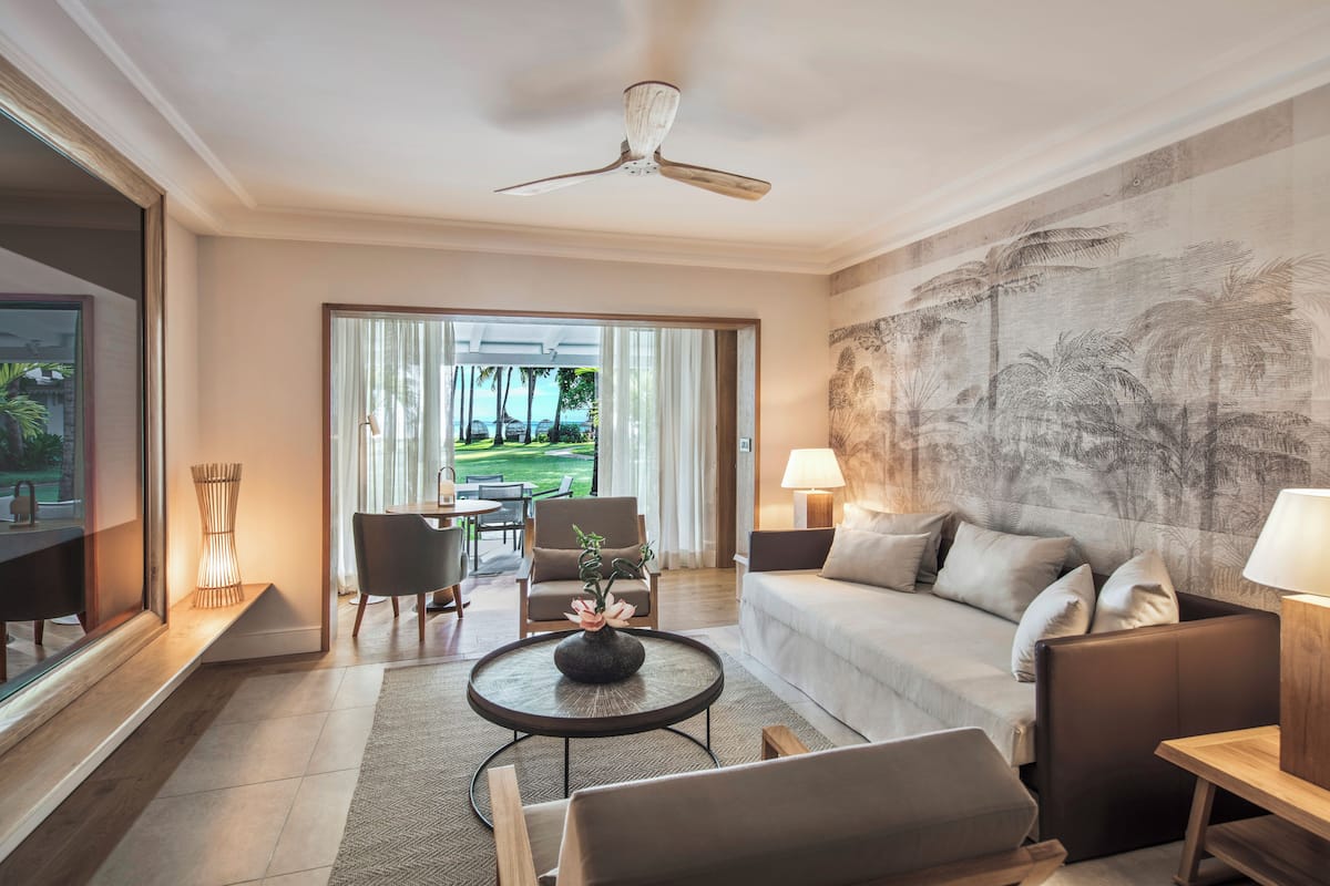 Una sala de estar moderna con sofá, sillón y mesa de centro. Hay un ventanal con vistas a un jardín y palmeras.