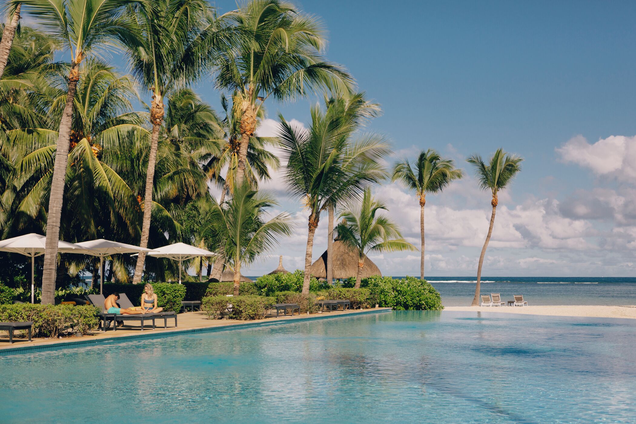 A poolside area with lounge chairs, palm trees, and a beach view.