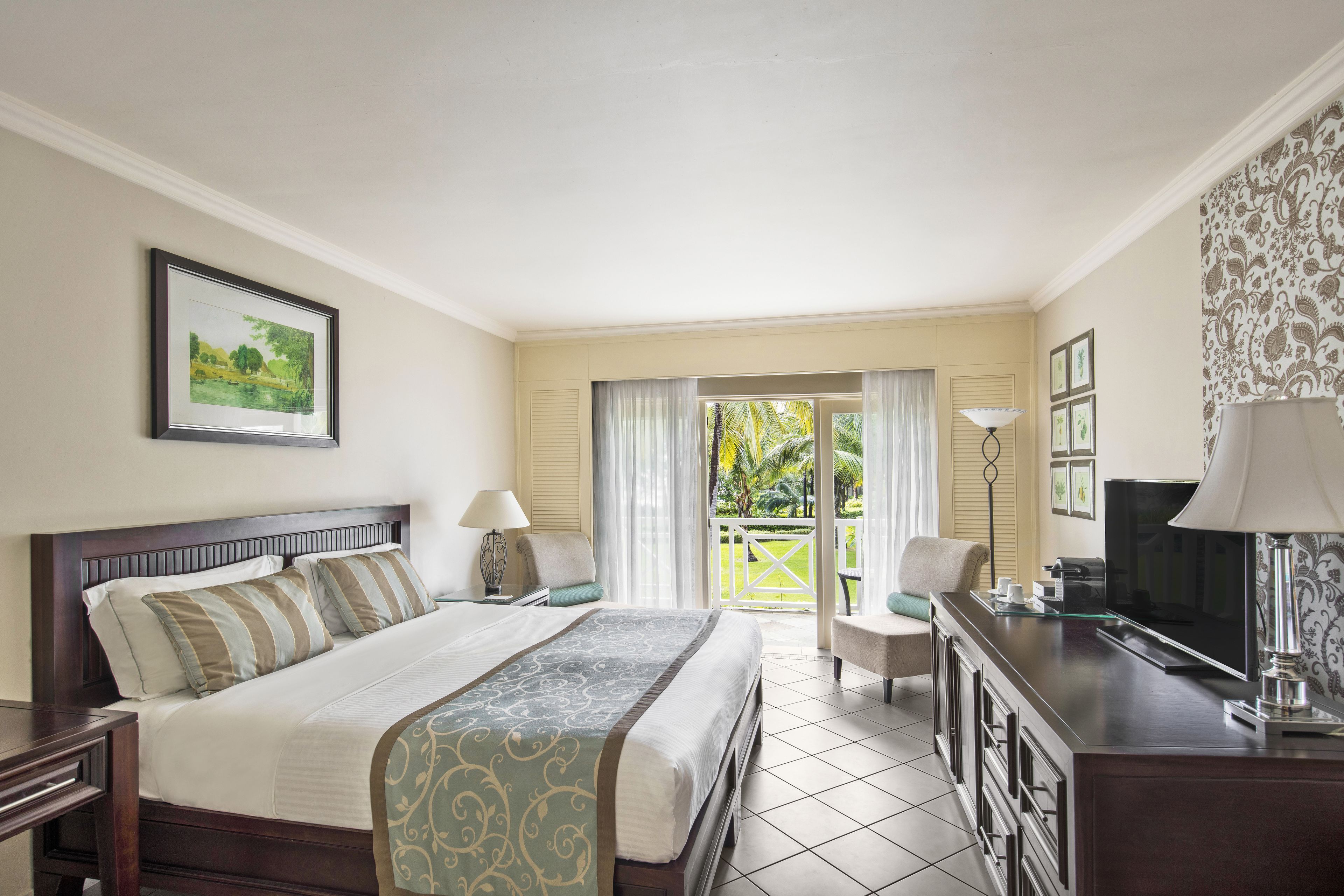 A hotel room with a large bed, a desk with a TV, a balcony with a view of palm trees, and a framed picture on the wall.