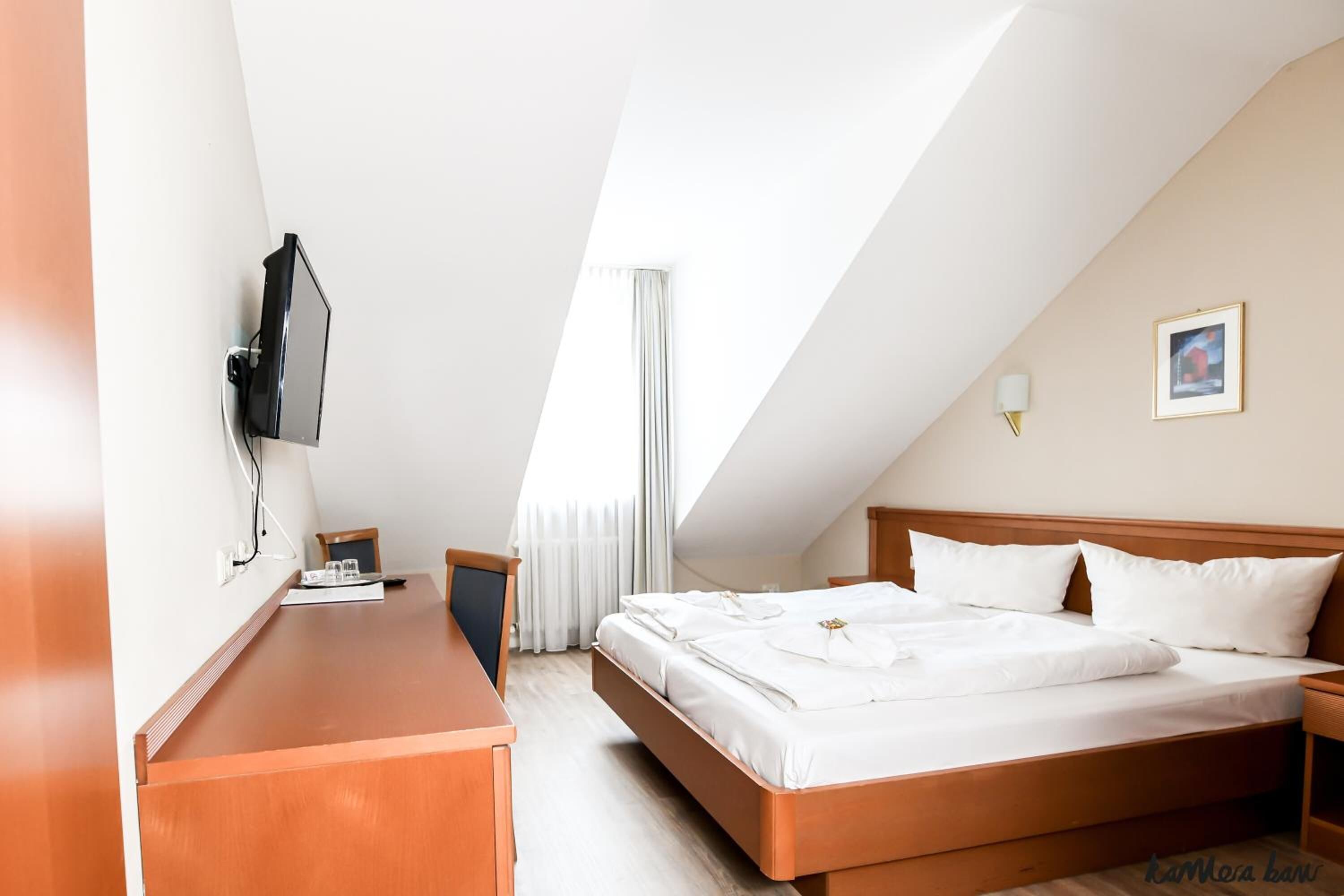 Une chambre d’hôtel avec un lit, un bureau, un téléviseur fixé au mur, une fenêtre avec des rideaux et un tableau encadré accroché au mur.
