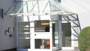 Entrada de hotel con fachada de vidrio, mostrador de recepción a la vista y un cartel con el nombre del hotel e información de contacto.