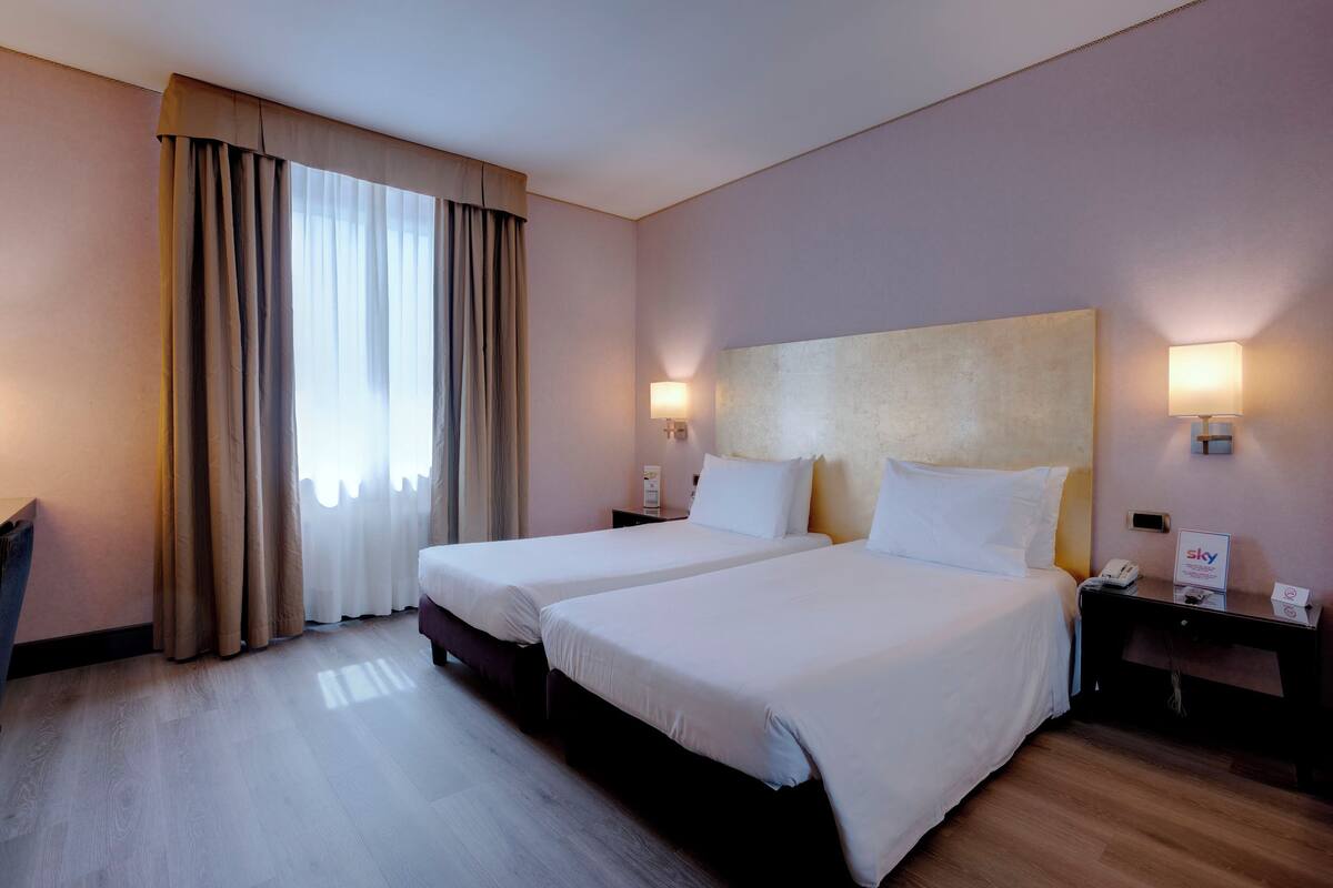 Ein Hotelzimmer mit zwei Betten, einem großen Fenster mit Vorhängen, Nachttischlampen und einem Nachttisch mit Telefon und Schild.