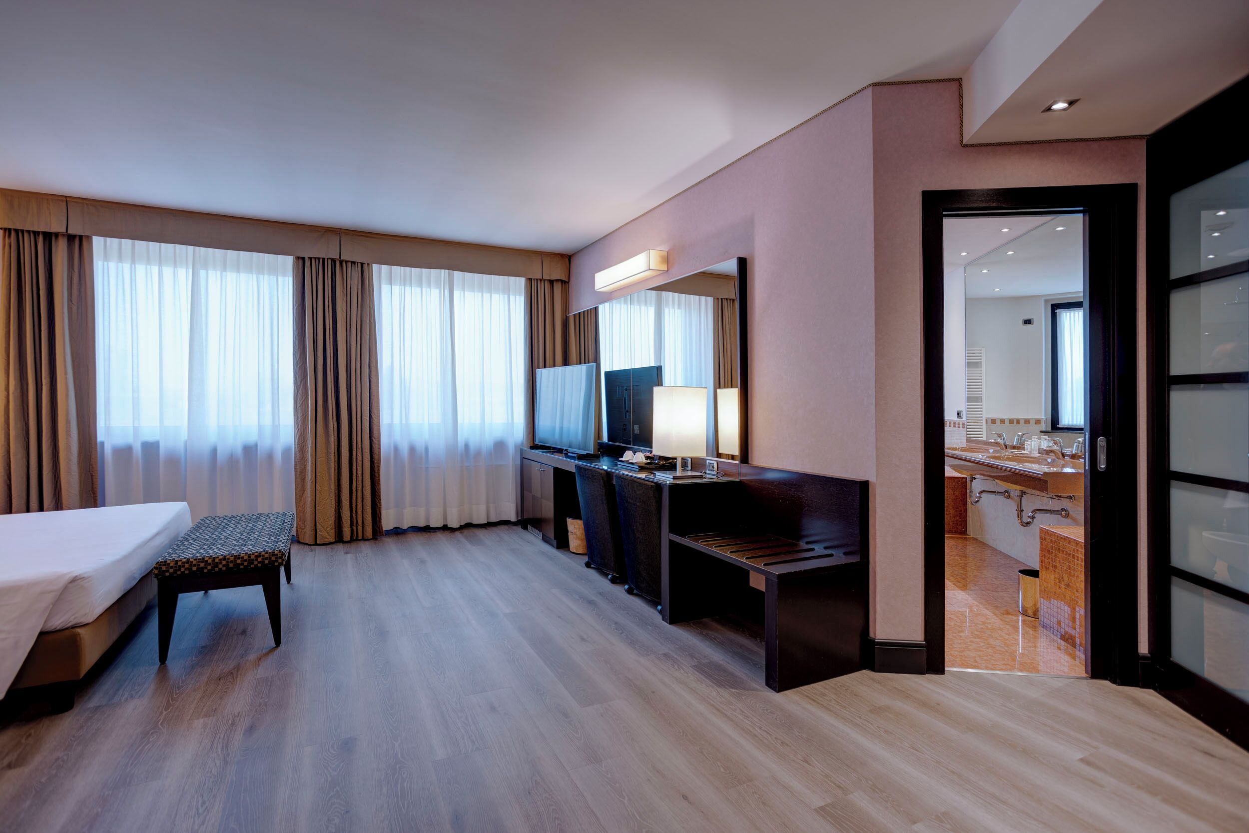 Habitación de hotel con cama, escritorio, silla, televisión y baño visible a través de una puerta abierta.