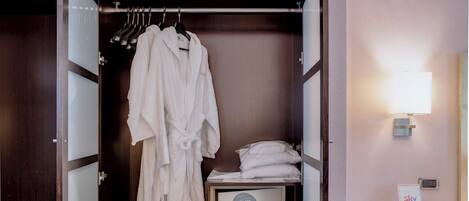 A walk-in closet with a hanging robe, a bed with pillows, a bedside table with a lamp, and a desk with a phone.