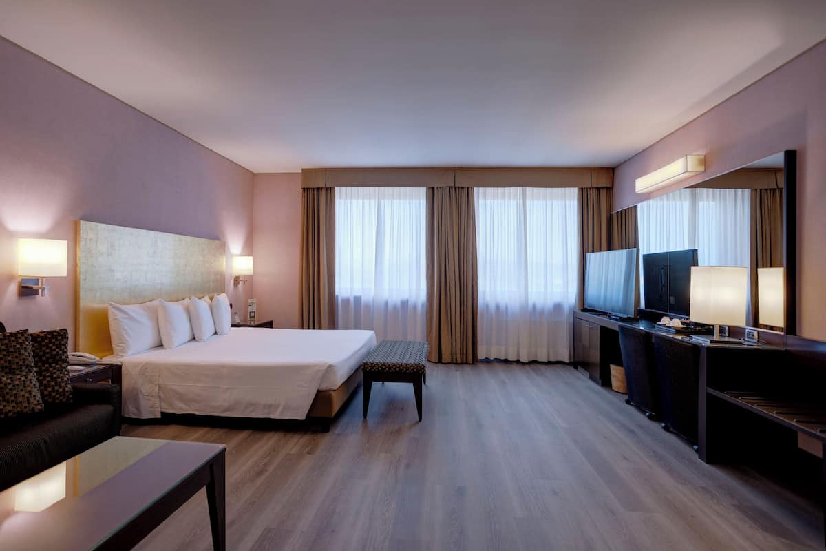 Ein Hotelzimmer mit einem Bett, einem Schreibtisch, einem Fernseher und einem großen Fenster mit Vorhängen.