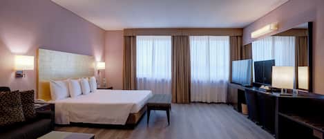A hotel room with a bed, a desk, a television, and a large window with curtains.