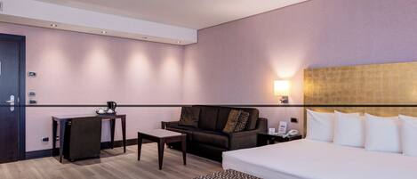A hotel room with a bed, a sofa, a small table, a bench, a desk, and a nightstand with a phone.
