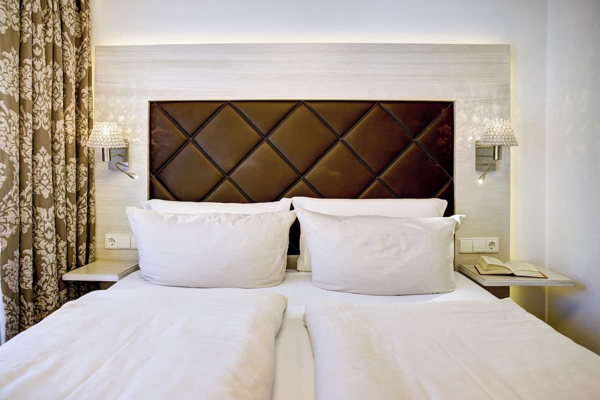 Uma cama de casal com cabeceira estofada, duas mesas de cabeceira com abajures e uma cortina com padrão floral.