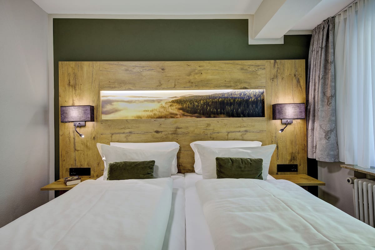 Uma cama de casal com lençóis brancos, duas luminárias de cabeceira, uma cabeceira de madeira e uma pintura de paisagem acima da cama.