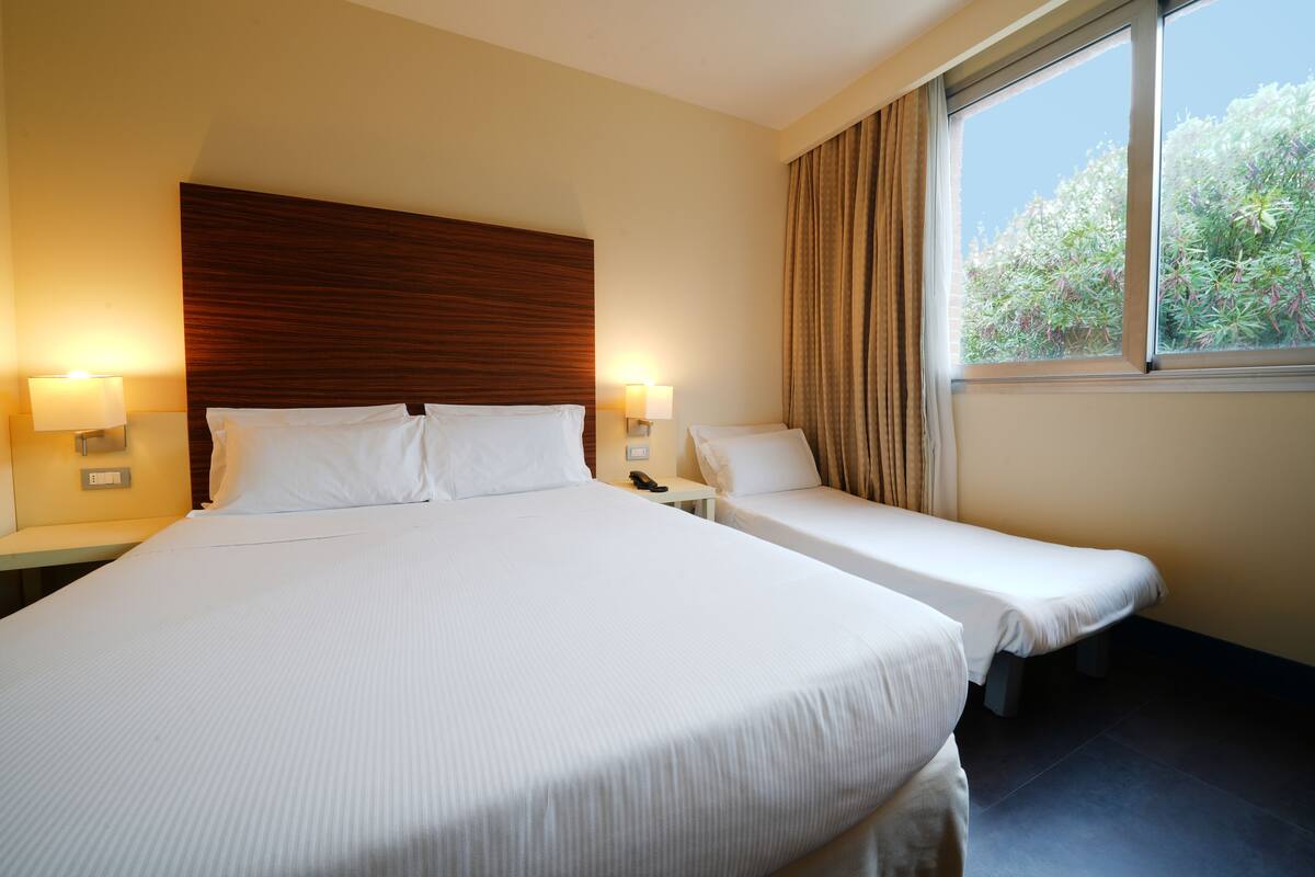 A hotel room with a large bed, two bedside tables, a wooden headboard, and a window with curtains.