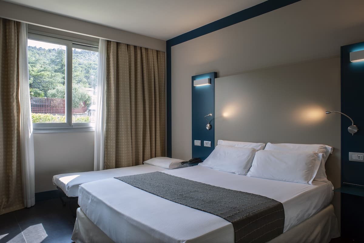 A hotel room with two beds, a large window with curtains, and a view of greenery outside.